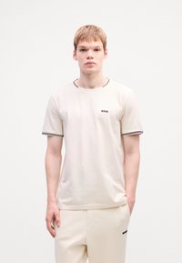 T-shirt crème à manches courtes avec un col rond, présentant des accents de rayures noires et un petit logo "BOSS" sur la poitrine. Texture de tissu douce.