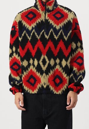 Sudadera de forro polar con cuello alto, que presenta un patrón geométrico en rojo, negro, crema y azul marino. Cierre de cremallera y puños elásticos.