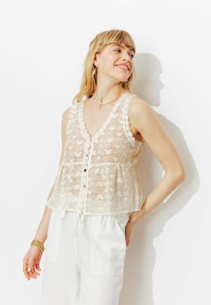 Femme souriante regardant vers la droite, portant un haut sans manches en dentelle blanche à boutons sur un pantalon blanc, accessoirisée d'un collier de perles et d'un bracelet en or.