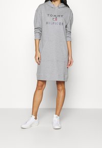 Vestido sudadera gris con capucha, que presenta un logo central "TOMMY HILFIGER" en letras oscuras y un bolsillo canguro en la parte frontal. Modelo con zapatillas blancas.