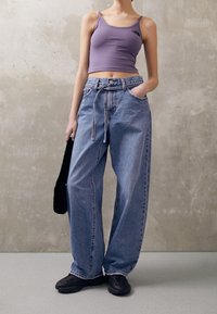 Fialový top s logem, světle modré volné džíny s vysokým pasem a černé tenisky. Modelka drží černou tašku a stojí na šedé podlaze.