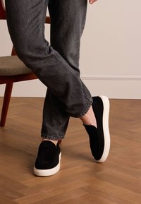 Black suede slip-on shoes with a white rubber sole, paired with dark gray cuffed jeans, against a wooden floor background.