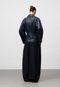 Donna con i capelli raccolti in un bun, che indossa una giacca di pelle blu navy oversize e pantaloni neri a gamba larga, in piedi di fronte a un muro grigio neutro.