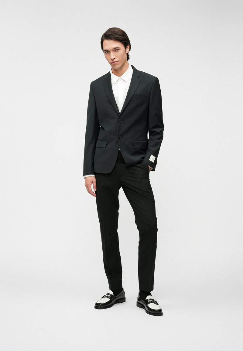 Schwarzer Anzug mit zwei Knöpfen, darunter ein weißes Hemd, schlanke Hose und schwarz-weiße Dressschuhe. Glattes Material, maßgeschneiderte Verarbeitung.