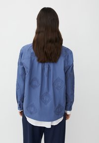 Osoba z długimi brązowymi włosami nosi niebieską koszulę z teksturowymi wzorami w diamenty, nakładaną na białą koszulę oraz ciemne spodnie, stojąc na białym tle.