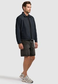 Mężczyzna ubrany w czarną kurtkę na zamek, czarne szorty dżinsowe oraz szare sneakersy New Balance, stoi na tle jednolitego, jasnoszarego tła.