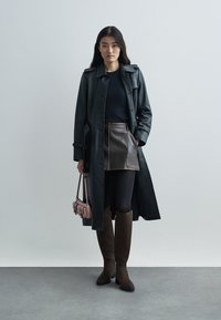 Manteau trench en cuir noir sur un haut bleu marine, associé à une mini-jupe en cuir marron et des leggings noirs. Bottes en daim marron jusqu'aux genoux, sac à main rose.