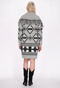 Halli hallitatud hall poncho mustade geomeetriliste mustritega, millel on frired näka ja keskmise pikkusega disain, koos põlvekõrguste mustade saapadega.