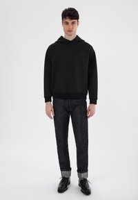Schwarzer Kapuzenpullover in lässiger Passform, kombiniert mit dunklen Jeans, hochgekrempelten Bündchen und schwarzen Schnürschuhen, vor einem hellen Hintergrund.