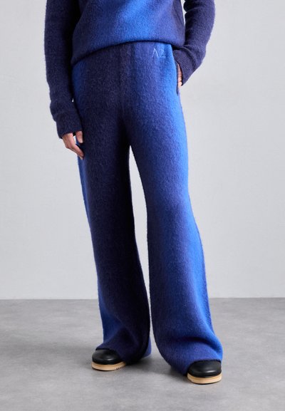 Blaue gradientfarbene Wollhosen mit einer weichen, strukturierten Oberfläche. Verfügt über ein weites Beindesign und ein gesticktes Logo am Bund. Schwarze Schuhe sind sichtbar.