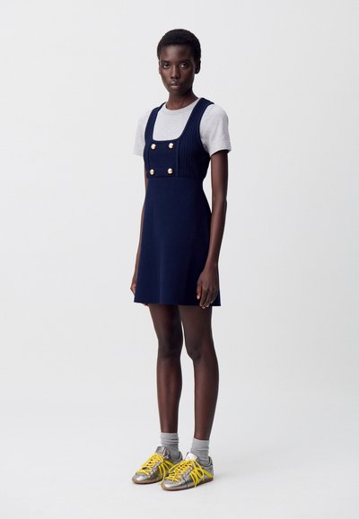 Robe en tricot navy avec des boutons dorés, présentant un décolleté carré. Associée à une chemise courte à manches grises clair et des baskets avec des accents jaunes.