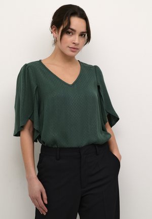 Blouse verte texturée avec un décolleté en V, des manches évasées et une coupe ample, assortie à un pantalon noir ajusté. Pas de motifs ni d'ornements visibles.