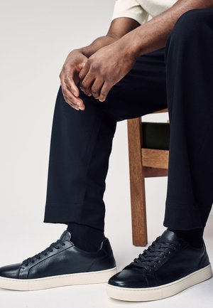 Zittend persoon draagt zwarte leren sneakers, zwarte sokken, zwarte broek en een wit kortarmig shirt, handen rustend op knieën.