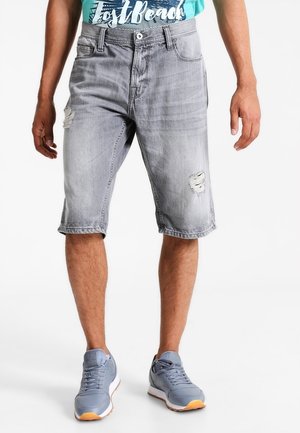 Mężczyzna noszący wyblakłe szare jeansowe szorty z wystrzępionymi łatami, w połączeniu z jasnoniebieskimi sneakersami i turkusowym t-shirtem z grafiką.
