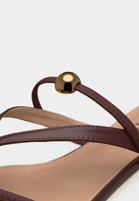 Καφέ σανδάλι με λεπτά λουράκια που διασταυρώνονται πάνω από το πόδι, με ένα μικρό στρογγυλό χρυσό μεταλλικό διακοσμητικό χάντρα σε ένα λουράκι.