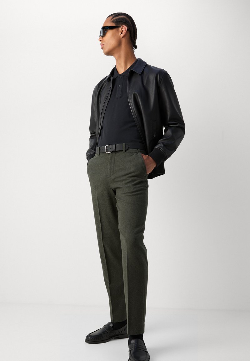 Schwarze Lederjacke, schwarzes Polo, olivgrüne Hose, schwarzer Gürtel und Slipper. Die Accessoires umfassen dunkle Sonnenbrillen. Das Modell steht selbstbewusst.