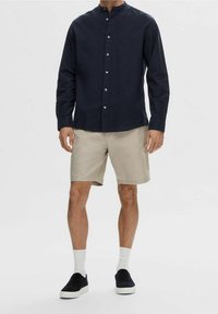 Chemise à manches longues bleu marine à boutons, short beige, chaussettes blanches et chaussures noires à enfiler ; tenue décontractée au coupe ample et au design simple.