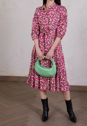 Femme portant une robe midi fleurie rose et crème avec des manches bouffantes, tenant un petit sac à main vert, et des bottines noires sur un sol en bois.