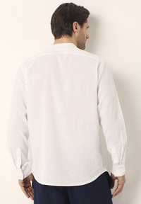 Camisa de lino blanca de manga larga con dobladillo recto, puños abotonados y textura suave; presenta costuras mínimas y un pequeño cuello.