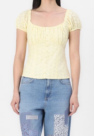 Žena nosící světle žlutou halenku s krátkými rukávy s děrováním, zapínanou na knoflíky vpředu, a modré džíny s našitými vzory.