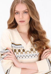 Kremowy sweter z dekoltem w kształcie litery V, z geometrycznym wzorem w brązie, niebieskim i żółtym na klatce piersiowej i rękawach. Miękka tekstura.