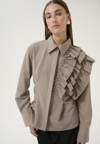 Blusa beige con colletto strutturato, chiusura a bottoni e un dettaglio di rouches laterale. Maniche lunghe e tessuto liscio con una leggera lucentezza.