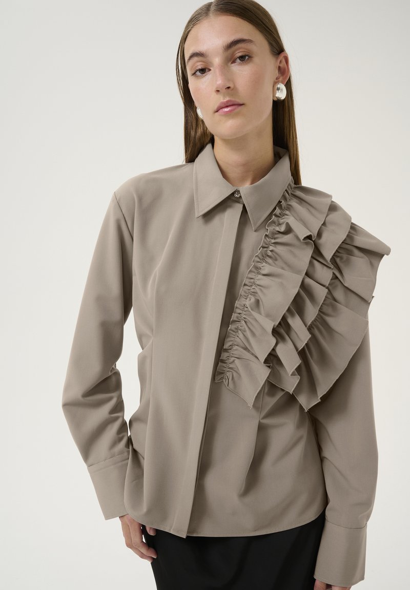 Blusa beige con colletto strutturato, chiusura a bottoni e un dettaglio di rouches laterale. Maniche lunghe e tessuto liscio con una leggera lucentezza.