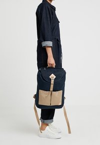 Granatowy plecak z klapą, brązowe przednie kieszenie, regulowane paski i diamentowy akcent na klapie.