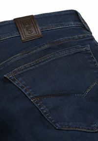 Jean en denim bleu foncé avec une surface texturée, une étiquette en cuir embossée avec le label "M5", et une poche arrière avec des coutures courbées.