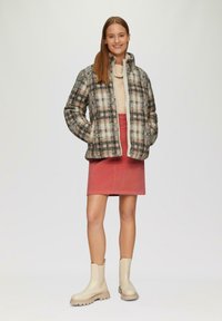 Doudoune à carreaux en beige et noir avec un col montant, portée sur un pull crème et une jupe rose, associée à des bottines beiges.