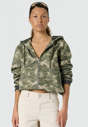 Kobieta nosząca okulary przeciwsłoneczne na głowie, zieloną bluzę z kapturem na suwak w kamuflażu oraz jasnobeżowe spodnie, stojąca na jednolitym tle.