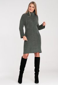 Strickkleid in Dunkelgrün, mit Rollkragen, langen Ärmeln, strukturiertem Stoff und asymmetrischem Saum. Kombiniert mit schwarzen kniehohen Stiefeln.