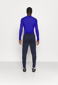 Urheiluasu, johon kuuluu istuva sininen pitkähihainen paita ja tummansiniset verkkarit; molemmat valmistettu sileästä, joustavasta kankaasta. Mukana mustat kengät.