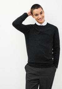 Čierny sveter s okrúhlym výstrihom, textúrovaná látka, úzky strih, kombinovaný so sivými nohavicami. Model má krátke vlasy a jemný úsmev.