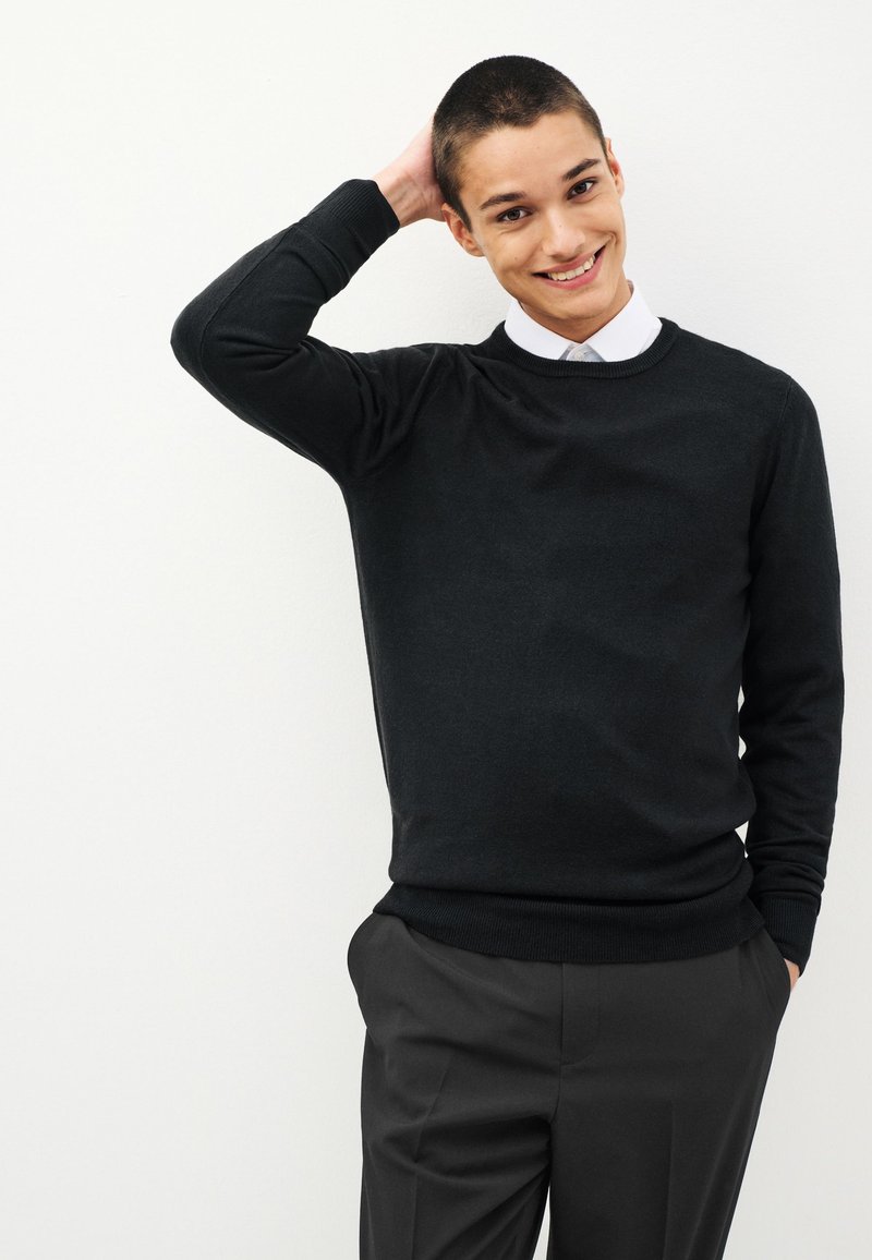 Čierny sveter s okrúhlym výstrihom, textúrovaná látka, úzky strih, kombinovaný so sivými nohavicami. Model má krátke vlasy a jemný úsmev.