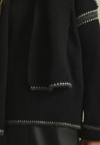 Fetter schwarzer Strickpullover mit geripptem Muster, der graue und weiße Strickakzente entlang der Ränder und Ärmel aufweist. Weiches, kuscheliges Material.
