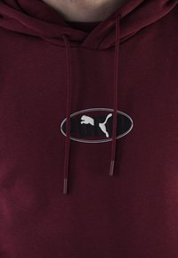 Sudadera color burdeos con capucha ajustable y un logo circular que presenta un gato saltando en blanco. Textura suave y diseño casual.