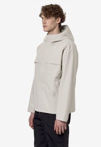 Giacca impermeabile grigio chiaro con cappuccio, due tasche frontali e design minimalista. Realizzata in tessuto liscio e resistente.