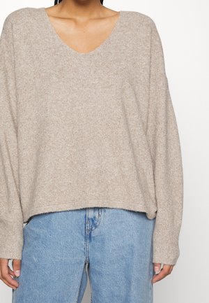Personne portant un pull en tricot beige ample avec un col en V et un jean taille haute bleu clair sur un fond uni.