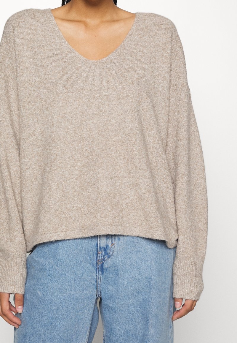 Personne portant un pull en tricot beige ample avec un col en V et un jean taille haute bleu clair sur un fond uni.