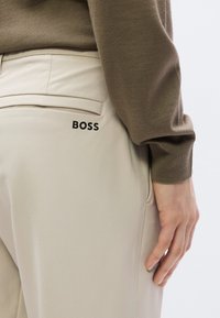 Μπεζ παντελόνια με πίσω τσέπη που φέρει μαύρο λογότυπο «BOSS». Το ύφασμα φαίνεται ομαλό και από πάνω  φοριέται ένα καφέ πουλόβερ.