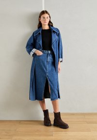 Trench coat in denim con collo oversize, camicia nera e gonna in denim al ginocchio con spacco anteriore. Stivaletti marroni con finitura testurizzata.