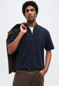 Homme aux cheveux courts et bouclés portant un polo bleu marine à col zippé et un pantalon marron, tenant une veste marron sur l'épaule, avec une expression neutre.