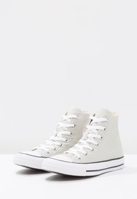 Baskets montantes en toile gris clair avec lacets blancs, bout en caoutchouc, bande noire le long de la semelle et détail du logo sur le côté.