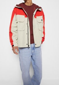 Kevyt beige-takki, jossa punaiset yksityiskohdat, kaksi etutaskua ja huppu. Yhdistetty viininpunaiseen paitaan ja vaaleansinisiin farkkuihin.