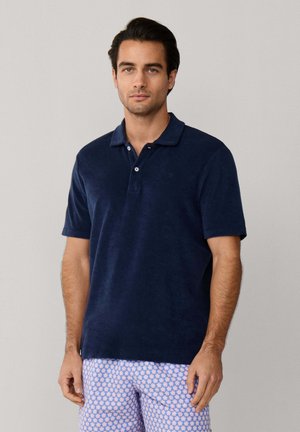 Mann trägt ein marineblaues Polohemd mit kurzen Ärmeln und hellblaue gemusterte Shorts, steht vor einem einfarbigen grauen Hintergrund.