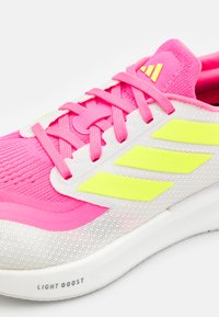 Baskets de sport roses et blanches avec une tige en maille respirante, présentant des accents jaunes néon et des lacets texturés. Logo de la marque visible.