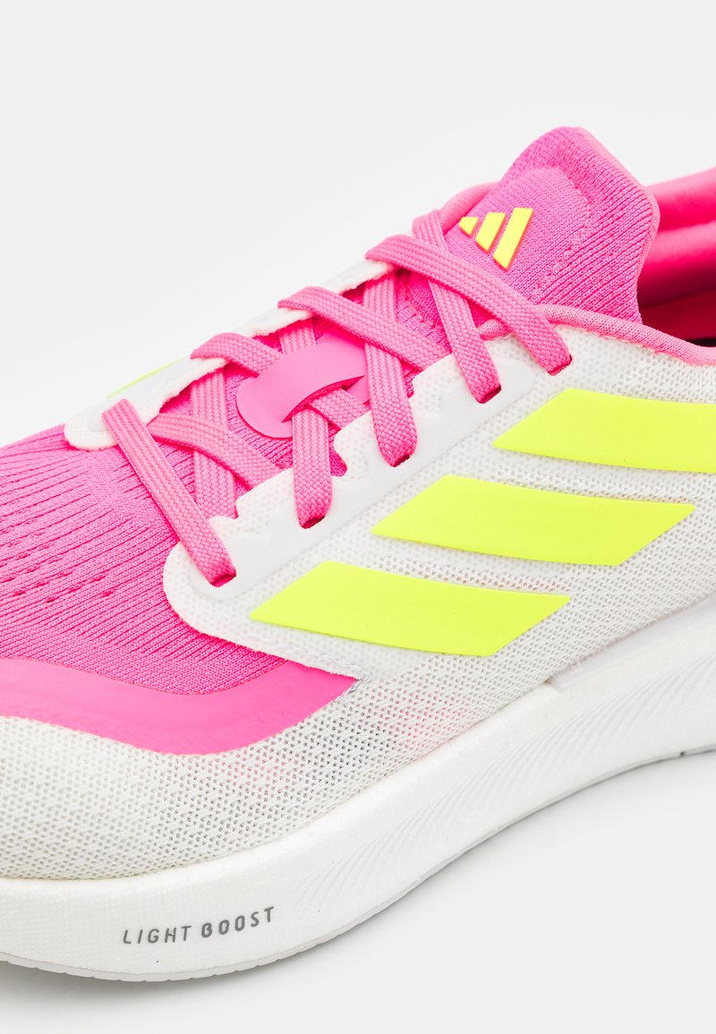 Zapatilla deportiva en rosa y blanco con parte superior de malla transpirable, con detalles en amarillo neón y cordones texturizados. Logo de la marca visible.