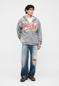 Νέος άνδρας με γκρι φούτερ με ροζ γκράφιτι γράμματα και ανοιχτό μπλε τζιν με μπαλώματα, στέκεται με τα χέρια πίσω από την πλάτη, κοιτάζοντας πλάγια.