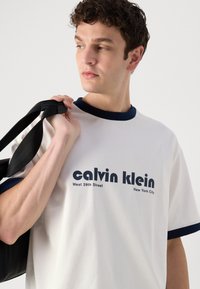 Hvit t-skjorte med marineblå krage og ermekanter. Har "calvin klein"-logoen og teksten "West 39th Street, New York City" i mørk skrift.